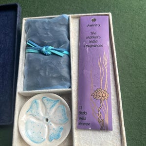 Op de afbeelding: Een cadeaupakket met wierook en een keramische wierookhouder. De set bevat een blauwe stoffen bundel met een lint, een witte en blauwe bloemen wierookhouder en een paarse verpakking met het opschrift "Amrita The Mother's India Fragrances" met 12 wierookstokjes.