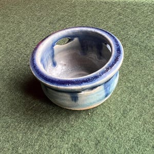 Può includere: Una ciotola in ceramica fatta a mano con un design unico. La ciotola presenta un'apertura circolare ed è dipinta con sfumature di blu e bianco. Il bordo è di un blu più scuro e la ciotola ha un aspetto strutturato. La ciotola viene probabilmente utilizzata per servire o decorare.