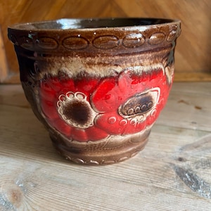 May include: A ceramic flower pot with a brown rim and a vibrant red floral design. The pot has a textured surface and a rounded shape, perfect for displaying plants. The pot is made of ceramic.