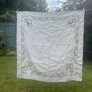 May include: A square white tablecloth featuring intricate cutwork embroidery along the borders. The design includes floral motifs in the corners and along the edges, finished with a scalloped edge detail throughout the entire perimeter.