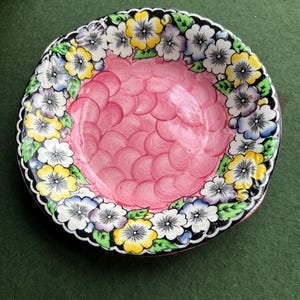 May include: A small pink saucer with a black rim and a floral design. The flowers are yellow, white, blue, and green.