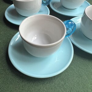 May include: A set of white ceramic teacups with light blue saucers. The teacups have a unique design with a transparent blue handle. The cups and saucers are arranged on a green surface.
