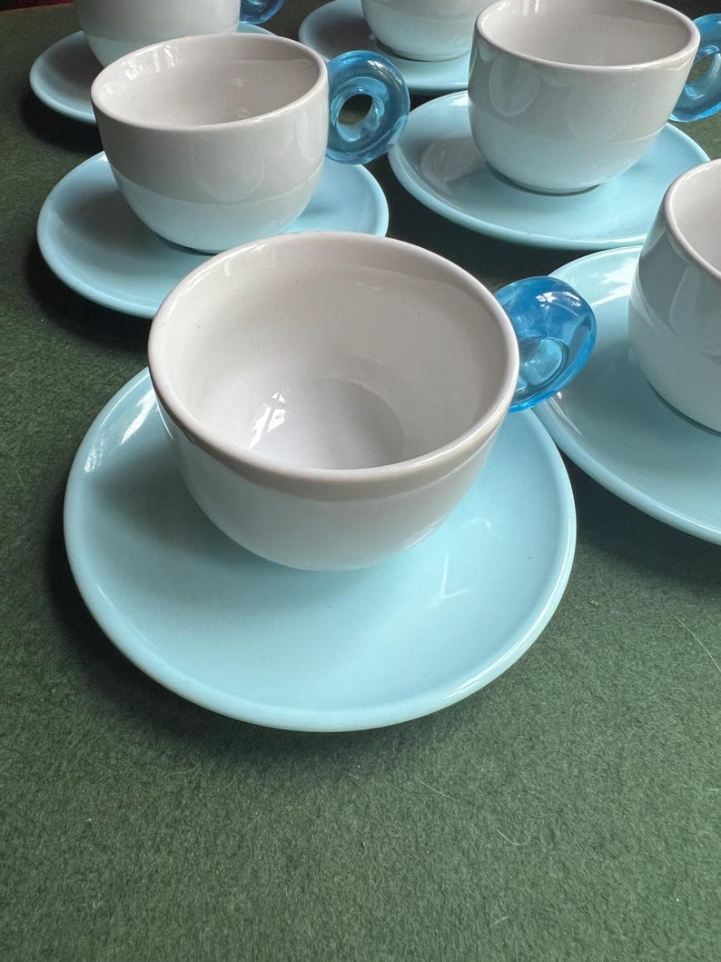 May include: A set of white ceramic teacups with light blue saucers. The teacups have a unique design with a transparent blue handle. The cups and saucers are arranged on a green surface.