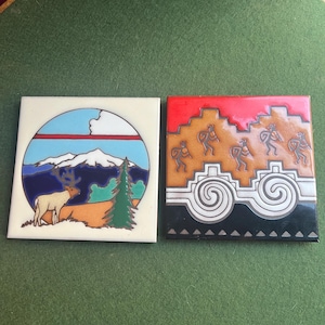 May include: Two square ceramic tiles with colorful designs. One features a mountain scene with a deer and pine tree. The other depicts figures and geometric patterns in red, brown, and black.