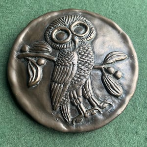 Op de afbeelding: Een bronzen, cirkelvormig reliëfsculptuur met een gedetailleerde uil die op een tak met bladeren zit. De uil heeft grote, expressieve ogen en ingewikkelde veerdetaillering. Het kunstwerk staat tegen een getextureerde achtergrond en toont een klassiek ontwerp.