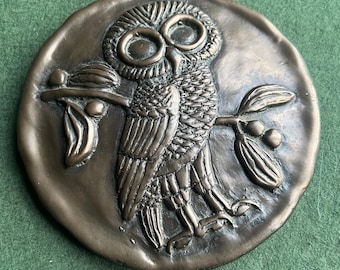 May include: A bronze-colored, circular relief sculpture featuring a detailed owl perched on a branch with leaves. The owl has large, expressive eyes and intricate feather detailing. The artwork is set against a textured background, showcasing a classical design.