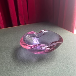 May include: A heart-shaped glass bowl, transitioning from pink to purple. The bowl is on a green surface, with a red curtain behind it. The glass has a smooth, glossy finish. The bowl is likely for decorative use.