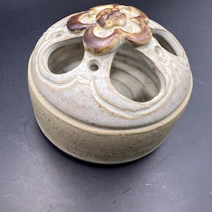 May include: A white ceramic pot with a brown flower design on the top. The pot has a round shape and is decorated with a swirl pattern. It has several holes around the top.