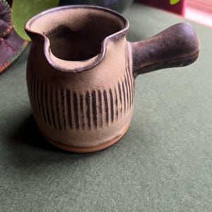 May include: A small, brown and tan ceramic creamer with a striped design and a handle. The creamer is sitting on a green surface.