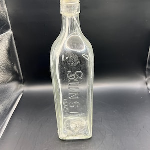 May include: Clear glass bottle with the embossed word "SUNST" and "T.C.P." on the front. The bottle has a rectangular shape with rounded shoulders and a narrow neck. The bottle is empty and has a light-coloured stopper.