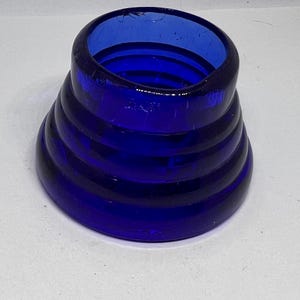 Puede incluir: Un objeto de vidrio azul cobalto con un dise&ntilde;o &uacute;nico y en capas. El objeto tiene una base ancha que se estrecha hacia arriba, con varias crestas horizontales que crean una apariencia apilada. La abertura superior es circular y el vidrio tiene un acabado brillante.