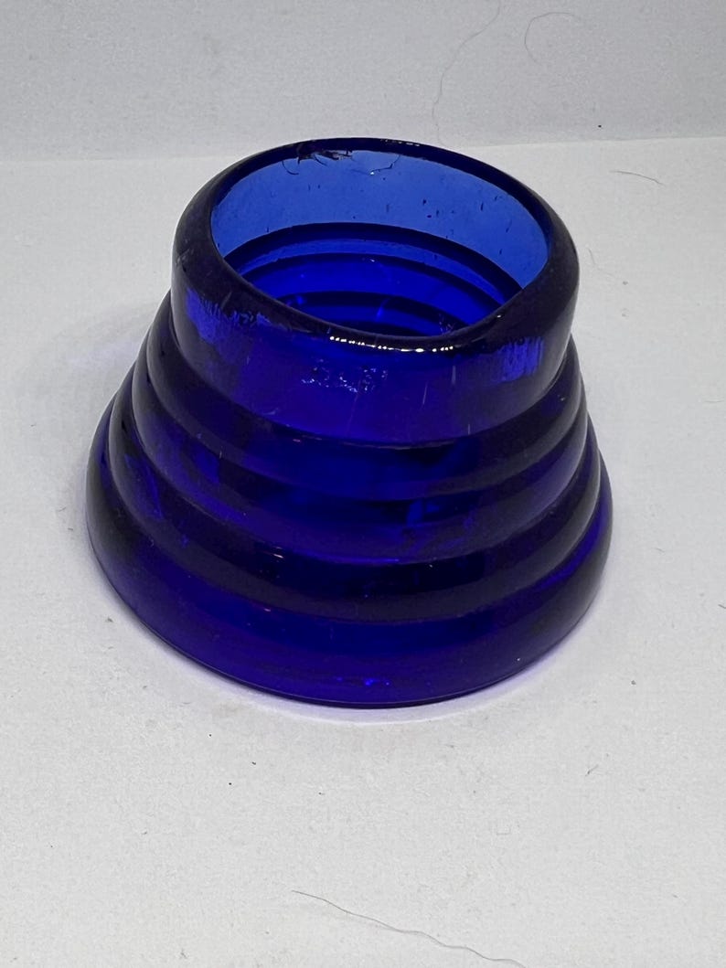 Puede incluir: Un objeto de vidrio azul cobalto con un dise&ntilde;o &uacute;nico y en capas. El objeto tiene una base ancha que se estrecha hacia arriba, con varias crestas horizontales que crean una apariencia apilada. La abertura superior es circular y el vidrio tiene un acabado brillante.