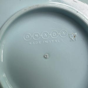 May include: A light blue ceramic plate with the text "OMADA" and "MADE IN ITALY" in white. The plate has a circular design with a slightly raised edge.