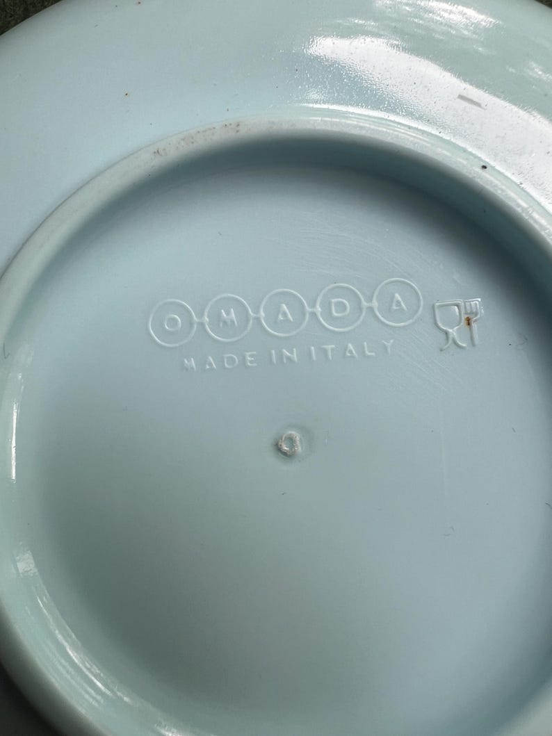 May include: A light blue ceramic plate with the text "OMADA" and "MADE IN ITALY" in white. The plate has a circular design with a slightly raised edge.