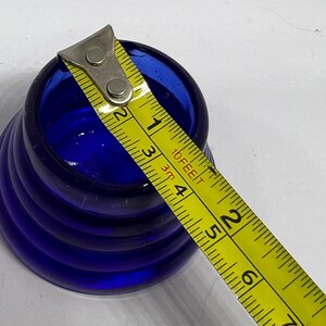 Puede incluir: Un objeto de vidrio azul cobalto con un exterior acanalado. Una cinta m&eacute;trica amarilla se coloca en la parte superior, mostrando medidas en cent&iacute;metros. El objeto parece ser un peque&ntilde;o jarr&oacute;n o recipiente.