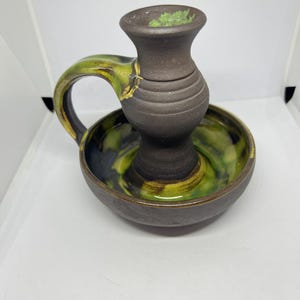 May include: A brown ceramic candle holder with a green and yellow glaze. The holder has a handle and a round base.