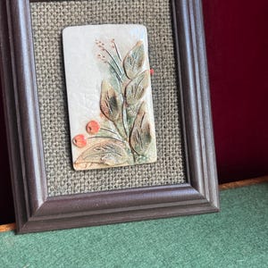 May include: A framed ceramic tile with a raised relief design of green leaves and red berries. The tile is set against a brown burlap background.