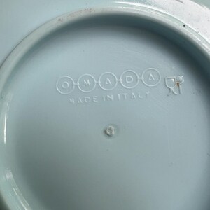 May include: A light blue ceramic plate with the text "OMADA" and "MADE IN ITALY" embossed on the bottom. The plate has a slightly raised rim and a small, circular indentation in the center.