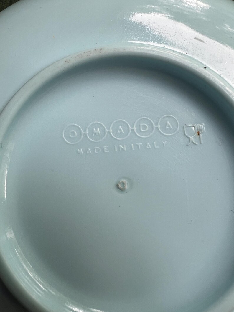 May include: A light blue ceramic plate with the text "OMADA" and "MADE IN ITALY" embossed on the bottom. The plate has a slightly raised rim and a small, circular indentation in the center.