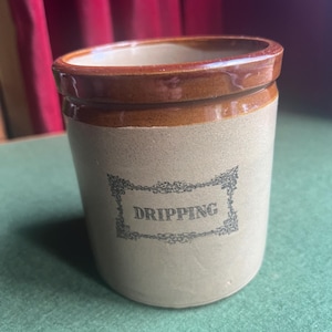 May include: A vintage stoneware jar with a brown rim and base, and a tan body. The word "DRIPPING" is printed in a decorative font within a rectangular frame on the front of the jar. The jar is cylindrical with a slightly tapered shape.