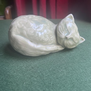 May include: A pale green ceramic figurine of a sleeping cat with a smooth, glossy finish. The cat is curled into a ball with its tail wrapped around its body, featuring subtle embossed floral patterns across its back and a peaceful facial expression.