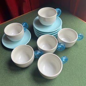 May include: A set of six white ceramic teacups with light blue handles and matching saucers. The cups are stacked on a green surface, showcasing a modern design. The handles are a vibrant blue.