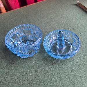 May include: Two light blue glass ring holders. One is a small bowl with a starburst pattern. The other has a central post for holding rings. Both are on a green surface.