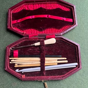 Puede incluir: Un estuche octogonal burdeos abierto, con un interior de terciopelo rojo, que contiene varios ganchillos y agujas. El estuche tiene un forro de terciopelo rojo y un ribete decorativo de cordón. Las herramientas incluyen ganchillos de madera, negros y blancos.