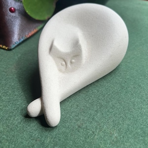 May include: White ceramic sculpture of a cat curled up sleeping. The cat's face is visible with closed eyes and a small nose.