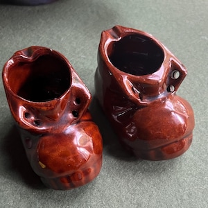 May include: Two brown ceramic boot-shaped ashtrays. The ashtrays are glazed and have a shiny finish.