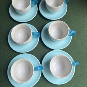 May include: Six white teacups with light blue saucers. Each cup has a transparent blue handle. The cups and saucers are arranged in two rows of three on a green surface. A classic tea set.