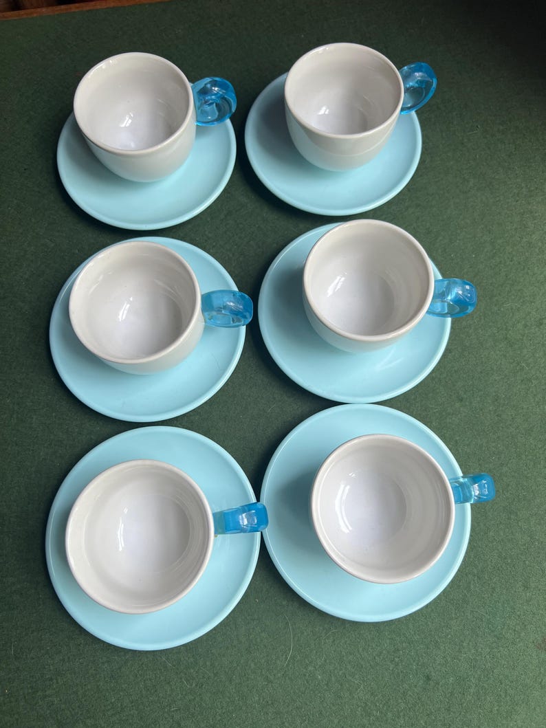 May include: Six white teacups with light blue saucers. Each cup has a transparent blue handle. The cups and saucers are arranged in two rows of three on a green surface. A classic tea set.