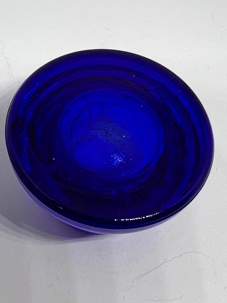 Puede incluir: Un cuenco de vidrio circular azul oscuro. El cuenco tiene una forma redondeada con una superficie lisa y brillante. El color es un azul rico y saturado, con sutiles variaciones de tono. El cuenco parece estar vac&iacute;o y est&aacute; sobre una superficie blanca.