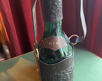 Puede incluir: Una botella de vidrio verde con un tapón metálico decorativo y un asa de transporte de cuero. La botella está encerrada en un soporte de cuero marrón con diseños florales en relieve. La palabra "GIN" está grabada en una etiqueta de cuero.