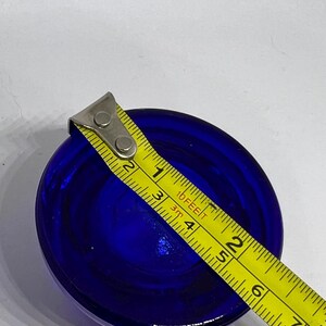 Puede incluir: Un objeto redondo de vidrio azul oscuro, de aproximadamente 7,6 cm de di&aacute;metro, se mide con una cinta m&eacute;trica amarilla. La cinta m&eacute;trica est&aacute; marcada en pies y pulgadas. El objeto tiene una superficie lisa y reflectante.