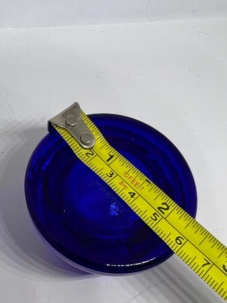 Puede incluir: Un objeto redondo de vidrio azul oscuro, de aproximadamente 7,6 cm de di&aacute;metro, se mide con una cinta m&eacute;trica amarilla. La cinta m&eacute;trica est&aacute; marcada en pies y pulgadas. El objeto tiene una superficie lisa y reflectante.