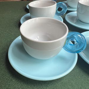 May include: A set of white ceramic teacups with light blue saucers. The teacups have a unique design with transparent blue glass handles. The cups and saucers are arranged on a green surface.