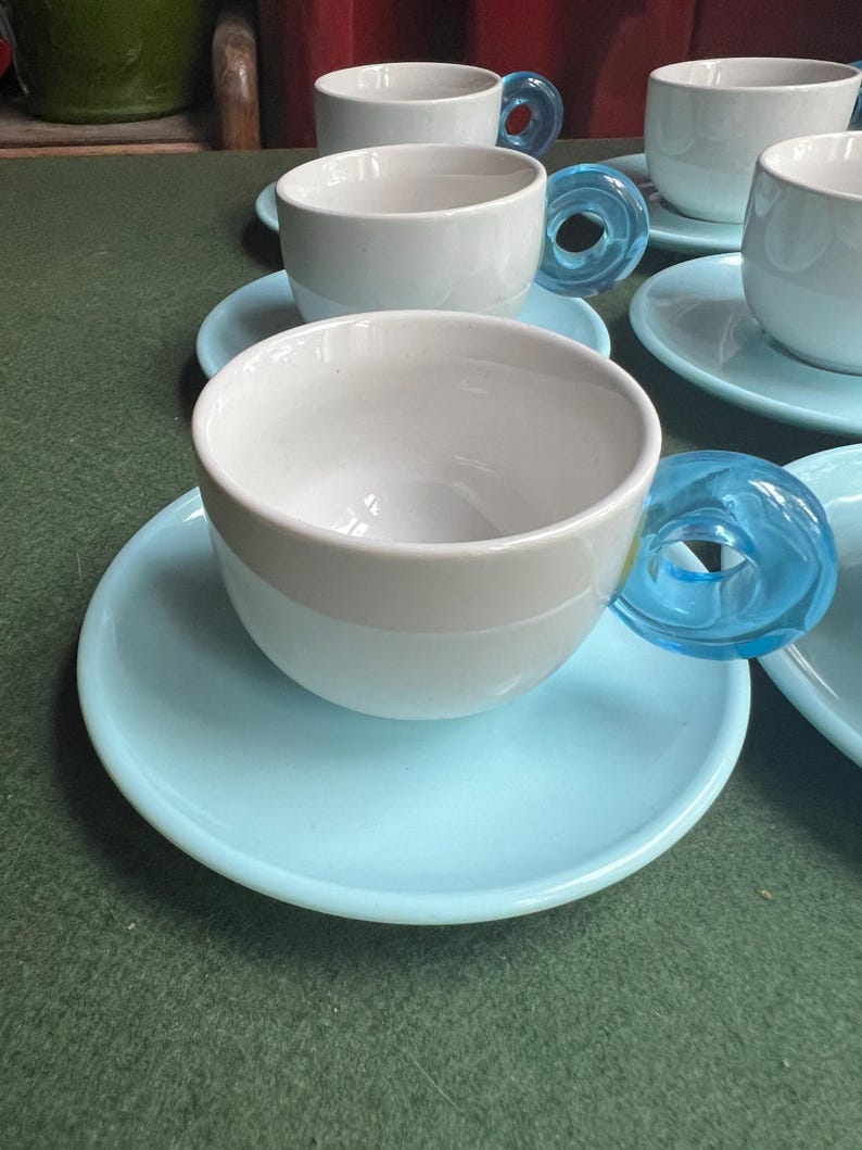 May include: A set of white ceramic teacups with light blue saucers. The teacups have a unique design with transparent blue glass handles. The cups and saucers are arranged on a green surface.