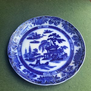May include: A round ceramic plate featuring a blue and white transferware pattern. The design depicts a landscape with pagodas, trees, and a small boat on water. The rim is decorated with an intricate floral and scroll border, and the surface has a visible fine crackle glaze finish throughout the entire piece.