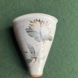 May include: A handmade, off-white ceramic wall pocket with a floral design. The pocket is cone-shaped with a wide opening and a narrow base. The flower is painted in shades of brown and blue-gray, with green leaves and stems. The pocket is speckled.