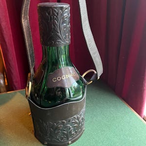 May include: A green glass Cognac bottle with a decorative metal holder and a leather strap. The bottle has a metal cap with an embossed floral design. The holder and cap are a dark bronze color. The word "COGNAC" is printed on the bottle.