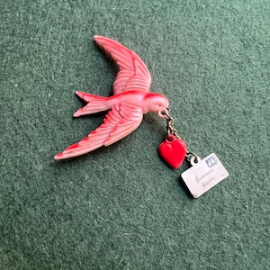 May include: A pink bird-shaped brooch with outstretched wings, holding a red heart and a small envelope charm. The bird has detailed wing and tail feathers. The heart and envelope are attached by a small chain.