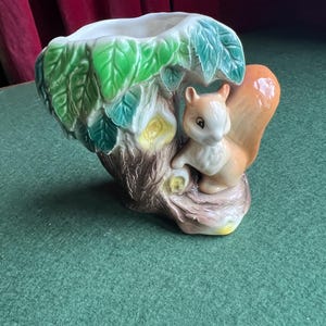 May include: A ceramic planter in the shape of a tree trunk with a squirrel. The planter has a white interior and is decorated with green leaves, brown bark, and an orange-tailed squirrel. The squirrel is perched on a branch.