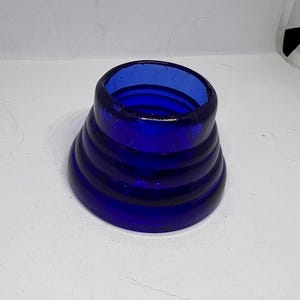 Puede incluir: Un objeto de vidrio azul cobalto con un dise&ntilde;o acampanado y acanalado. El objeto tiene una abertura ancha en la parte superior y se estrecha hacia una base m&aacute;s ancha. El vidrio parece liso y s&oacute;lido.