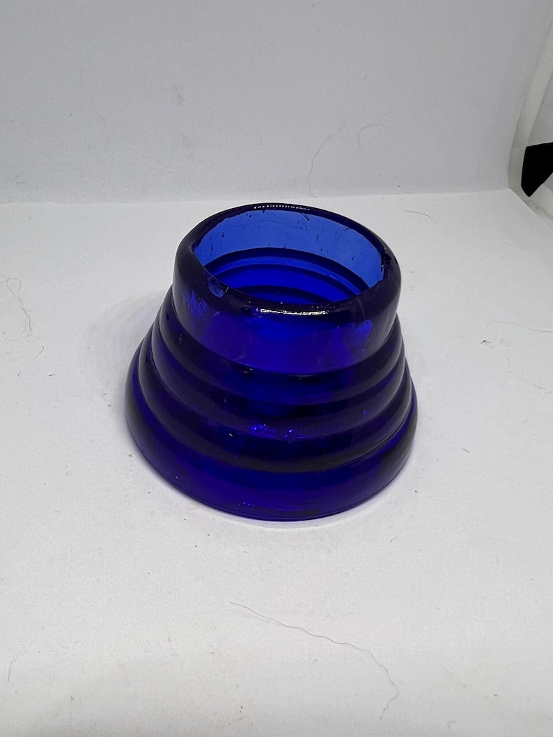 Puede incluir: Un objeto de vidrio azul cobalto con un dise&ntilde;o acampanado y acanalado. El objeto tiene una abertura ancha en la parte superior y se estrecha hacia una base m&aacute;s ancha. El vidrio parece liso y s&oacute;lido.