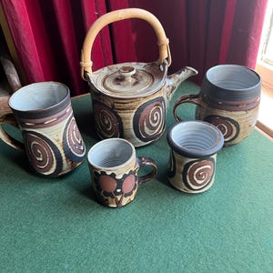 May include: A set of brown and white ceramic mugs and a teapot with a wooden handle. The mugs and teapot have a swirling pattern and are decorated with brown and white accents.