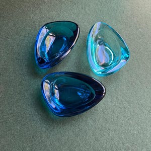 May include: Three glass ashtrays in shades of blue. The ashtrays have a unique, organic shape with rounded edges. The glass appears smooth and reflective, with a slight translucence. The ashtrays are arranged on a green surface.