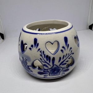 May include: A white ceramic candle holder with a blue and white floral design and cut-out hearts. The holder is round and has a small opening at the top for a candle.