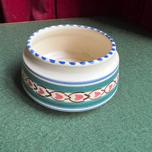 May include: A round ceramic bowl with a cream-colored interior and a decorative exterior. The rim has blue dots, and the body features a teal band with a repeating pattern of red hearts and brown accents. The bowl sits on a green surface.