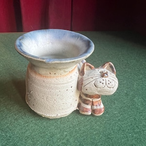 May include: A ceramic cat-shaped planter with a light beige body and a blue-rimmed opening. The cat has brown and white striped details and a smiling face. The planter is on a green surface.